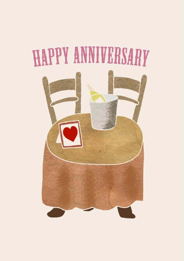 small round table covered with a tablecloth. There is an ice bucket and a bottle of champagne on the table. Also on the table is also a picture frame with an image of a red heart and text 'Happy Anniversary'